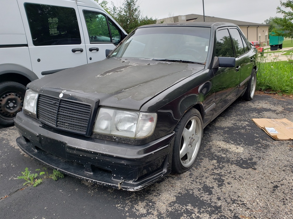 Front three quarter view of a 1986 Mercedes-Benz 300E with peeling paint job and flat tires.