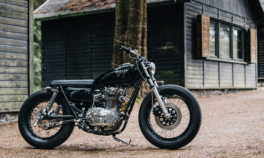 Right side shot of a 1974 XS650 painted black with silver suspension and engine sitting in front of a ,weathered shed.